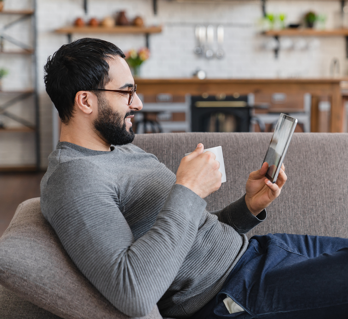 Een man zit op de bank met een kop koffie en een tablet in zijn hand.
